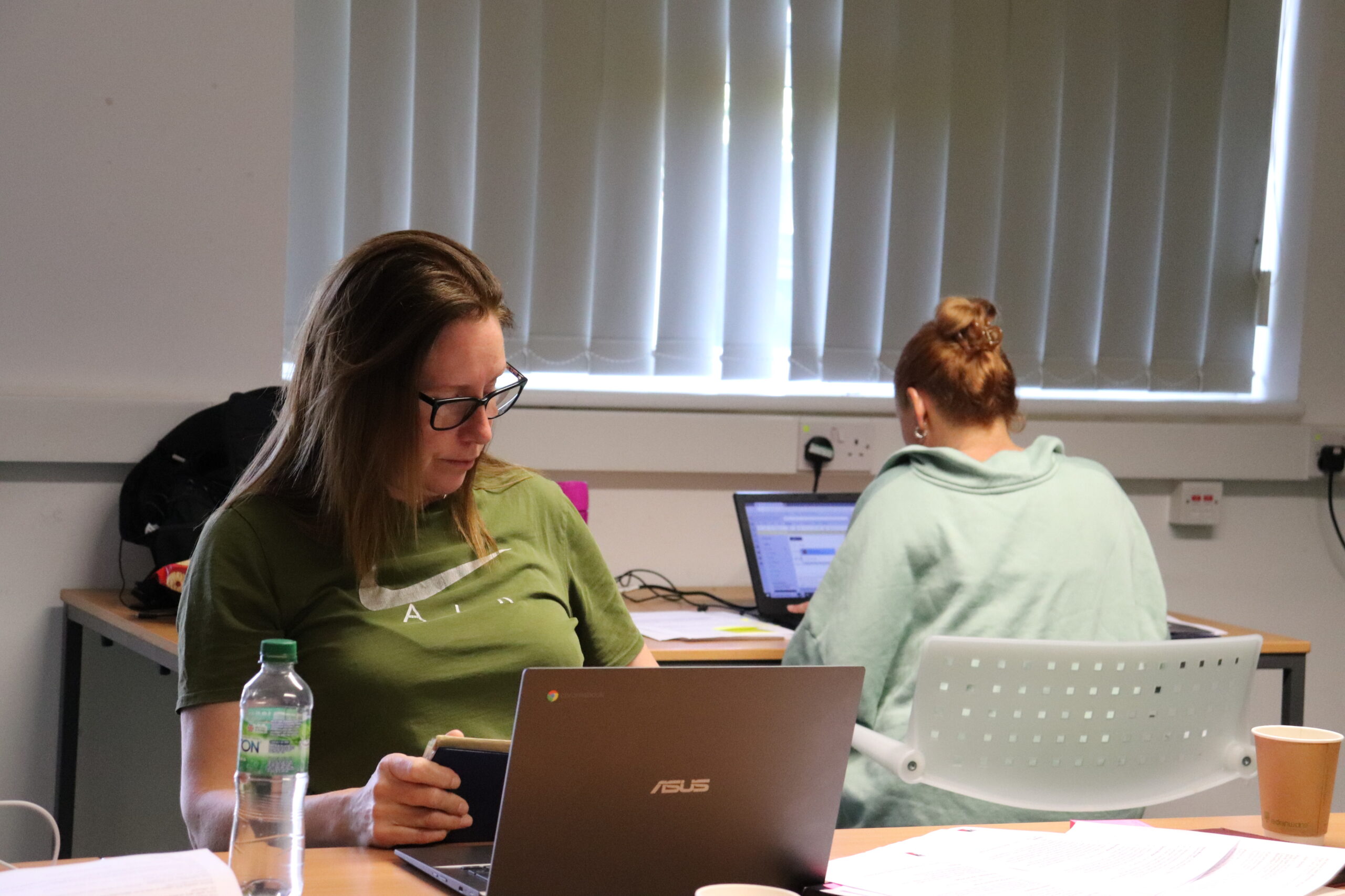 Two individuals are working at desks with laptops in a classroom setting.