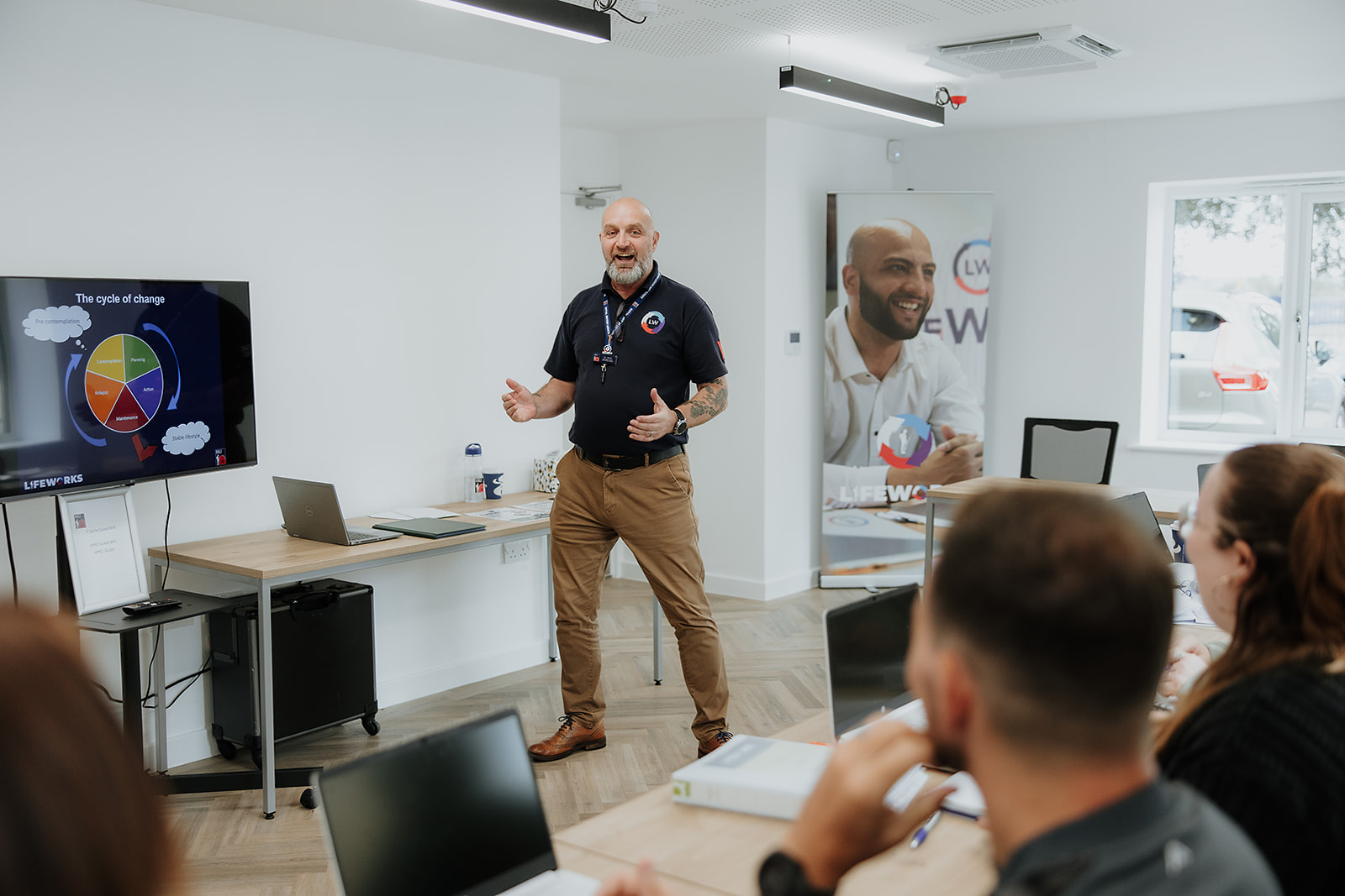 Two individuals are working at desks with laptops in a classroom setting.
