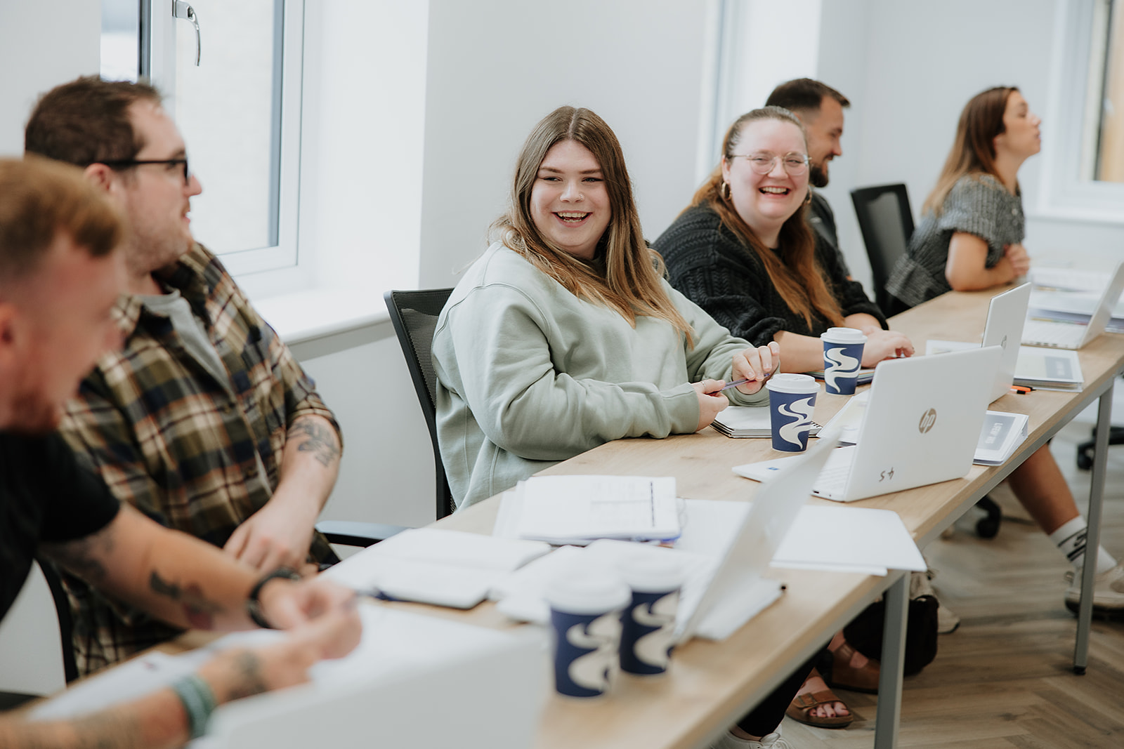 Two men are working together at a desk with laptops, one pointing at the other's screen. 