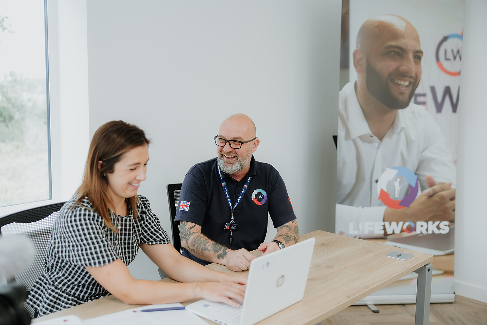 Two individuals are working at desks with laptops in a classroom setting.